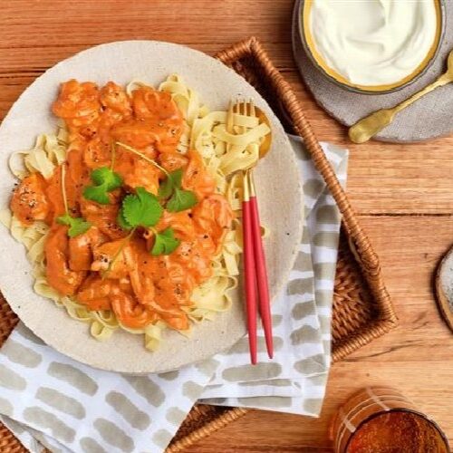 A plate of Butter Chicken Fettuccine on a woven tray next to beverage glasses and toppings.