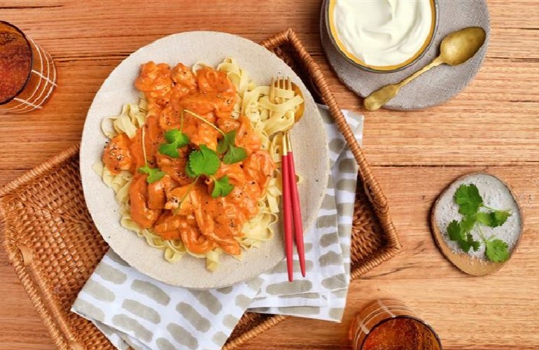 A plate of Butter Chicken Fettuccine on a woven tray next to beverage glasses and toppings.