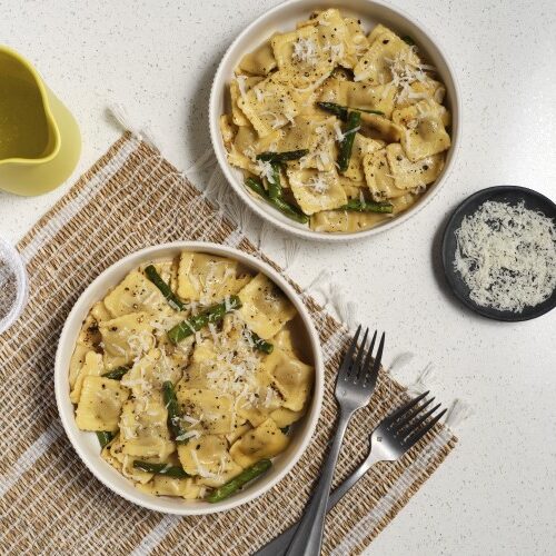 Two bowls of Cacio e Pepe Beef Ravioli with Asparagus next to utensils and beverages ready to serve for dinner