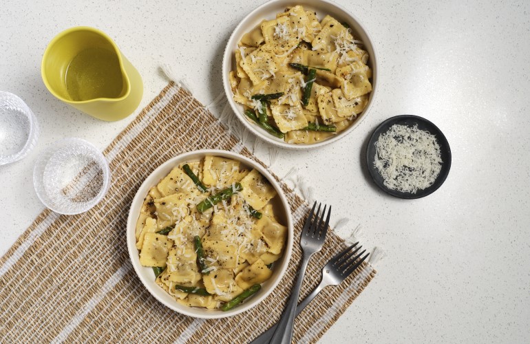 Two bowls of Cacio e Pepe Beef Ravioli with Asparagus next to utensils and beverages ready to serve for dinner