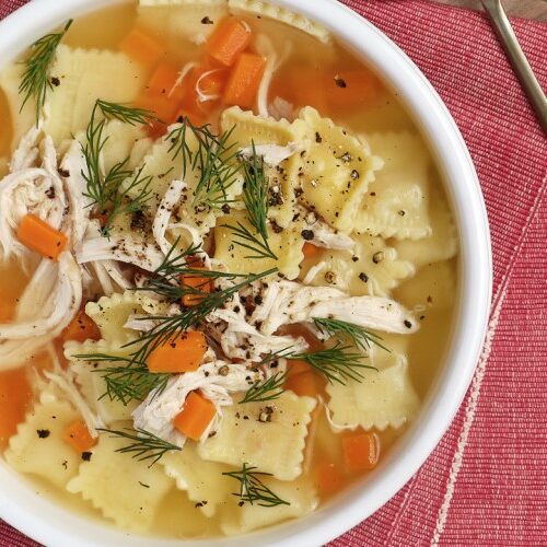 Bowl of Chicken Soup with Ravioli with red napkins and utensils on a wood serving board and wood surface.