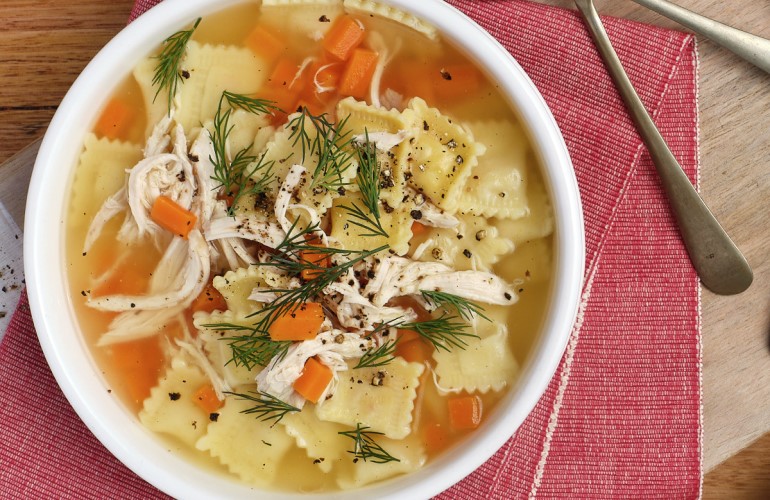 Bowl of Chicken Soup with Ravioli with red napkins and utensils on a wood serving board and wood surface.