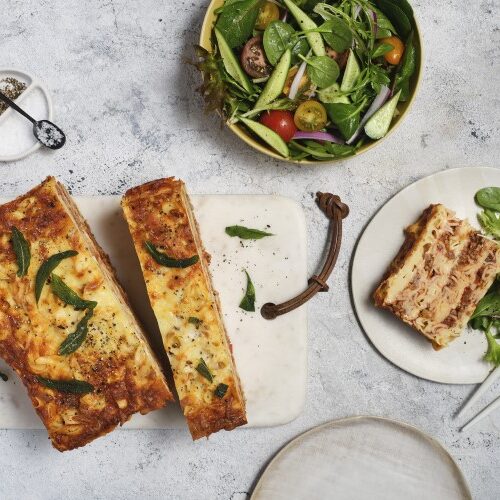 A Fettucine Bake cut into slices on a cutting board and a serving on a plate next to a bowl of salad greens.