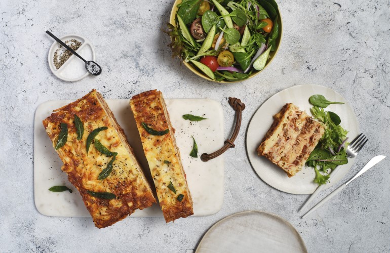 A Fettucine Bake cut into slices on a cutting board and a serving on a plate next to a bowl of salad greens.