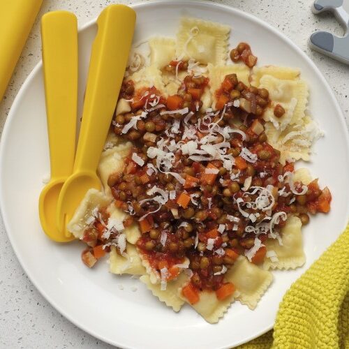 A white plate filled with Speedy Lentil Bolognese with a yellow child-size spoon and fork resting on the plate. A yellow napkin and fun wooden toy animals are also on the surface next to the plate.