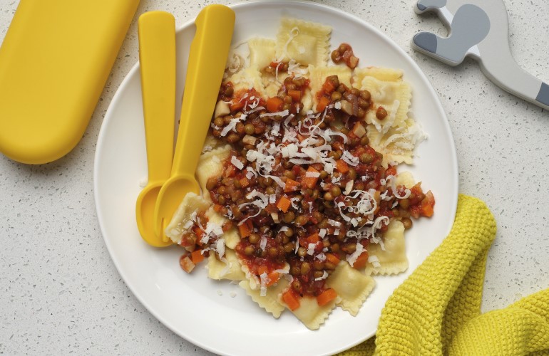 A white plate filled with Speedy Lentil Bolognese with a yellow child-size spoon and fork resting on the plate. A yellow napkin and fun wooden toy animals are also on the surface next to the plate.