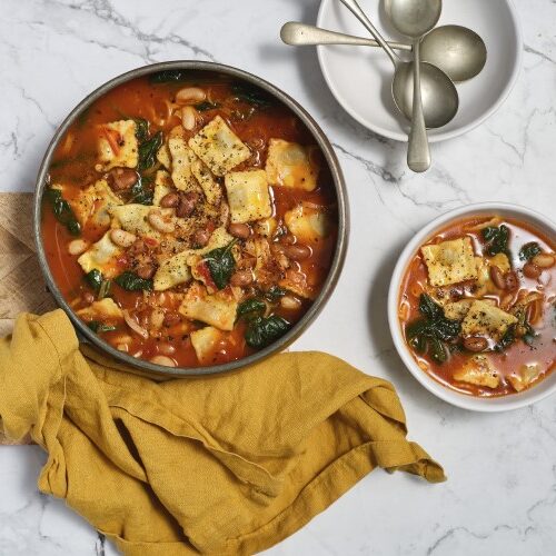 A bowl of soup, next to spoons, a small loaf of bread and a skillet of Tuscan Beef Ravioli Soup on a cutting board.