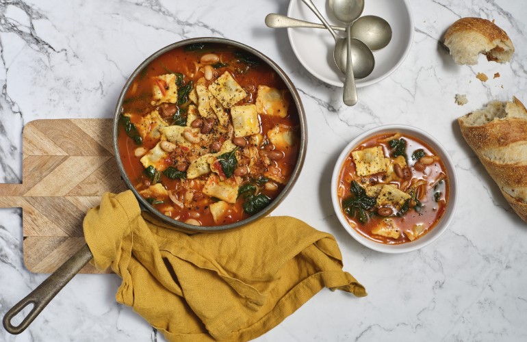 A bowl of soup, next to spoons, a small loaf of bread and a skillet of Tuscan Beef Ravioli Soup on a cutting board.